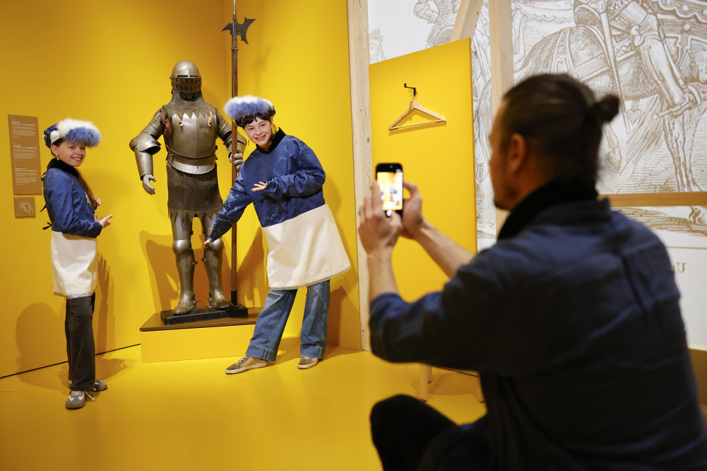 Dans une alcôve jaune vif d'un musée, deux jeunes filles déguisées en tuniques bleues et blanches, avec des chapeaux bordés de fourrure, posent en souriant de part et d'autre d'une armure médiévale complète tenant une hallebarde. Au premier plan, un homme accroupi les photographie avec son smartphone.