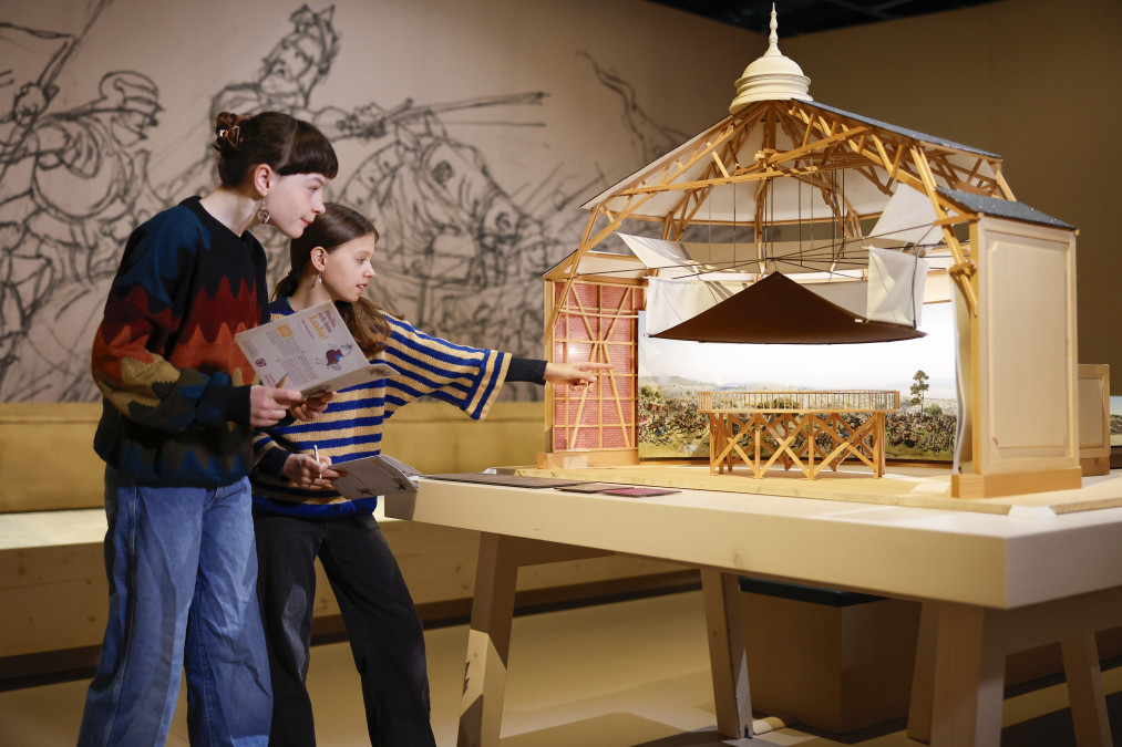 Deux jeunes filles observent une grande maquette en bois d'une rotonde de panorama, coupée pour montrer sa charpente, son dôme et, à l'intérieur, une plateforme d'observation entourée d'une peinture panoramique de bataille. L'une tient un livret d'activités, l'autre pointe la maquette du doigt. Au mur, un grand croquis au fusain de cavaliers en armes.