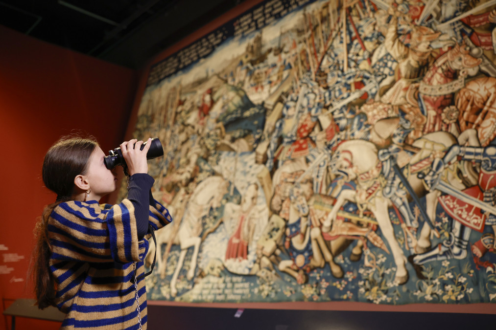 Une jeune fille en pull rayé bleu et beige regarde aux jumelles une immense tapisserie médiévale représentant une bataille : chevaliers en armure, chevaux, lances et épées, avec un bandeau de texte en haut et des fleurs en bas. La tapisserie est exposée sur un mur rouge.