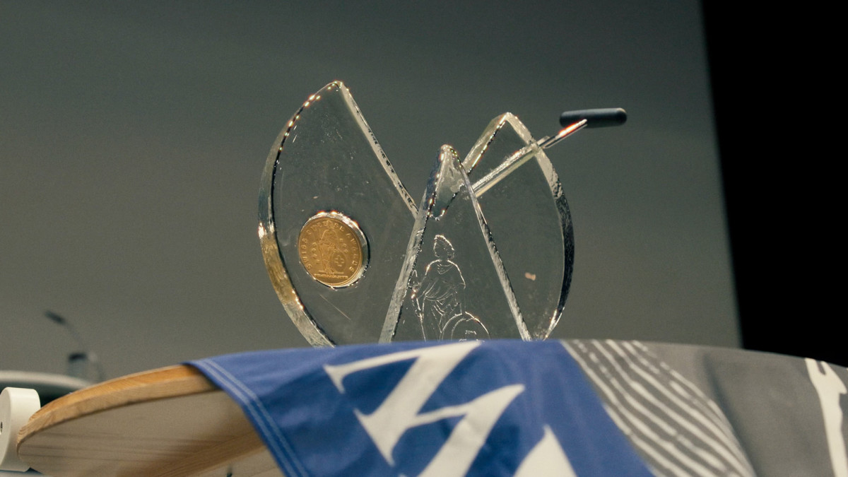 alt="Trophée en verre gravé des Swiss Cheese Awards, orné d'une pièce d'or, posé devant un drapeau bleu lors d'une cérémonie
